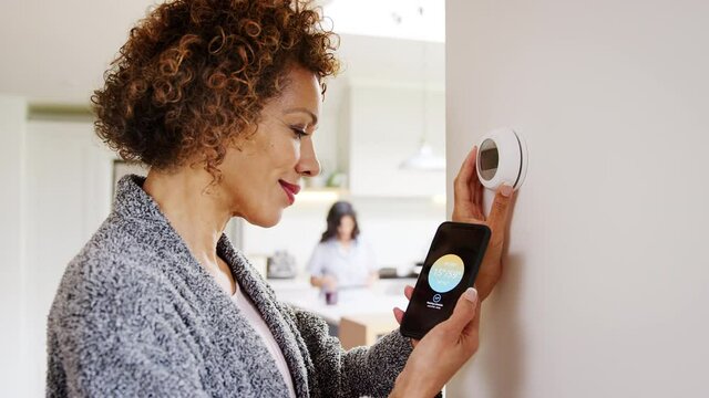 Mature woman using app on phone to control digital central heating thermostat at home with gay partner in background - shot in slow motion