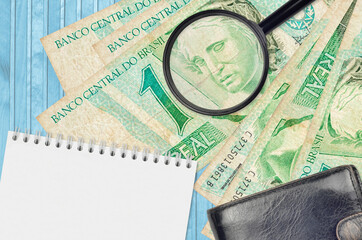 1 Brazilian real bills and magnifying glass with black purse and notepad. Concept of counterfeit money. Search for differences in details on money bills to detect fake