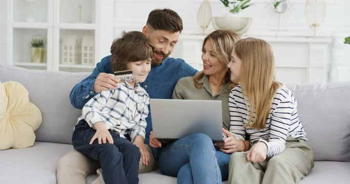 Caucasian mother and father shopping online with son and daughter on computer. Parents using credit card and paying in iternet on laptop. Family with kids spending money together on couch at home.