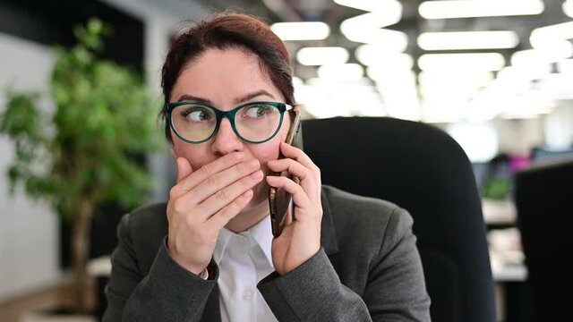 A woman sits in an office at her desk and gossips on the phone. Corporate ethics. Female employee in a suit tells secrets on a smartphone at work.