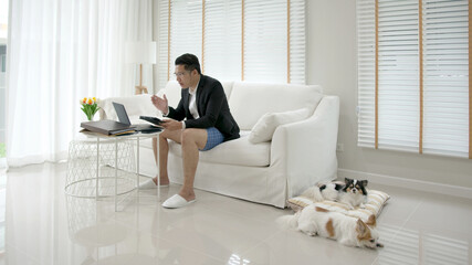 Asian man wearing a suit or business wear on top and sweatpants or boxers on bottom. Businessman video conference using laptop and tablet online meeting.Working from home and Working remotely.