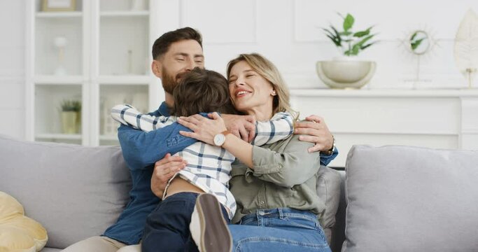 Rear of Caucasian cute little kid boy running to young happy mother and father sitting on couch in cozy living room. Cheerful parents hugging and embracing their small son at home on sofa. Back view.