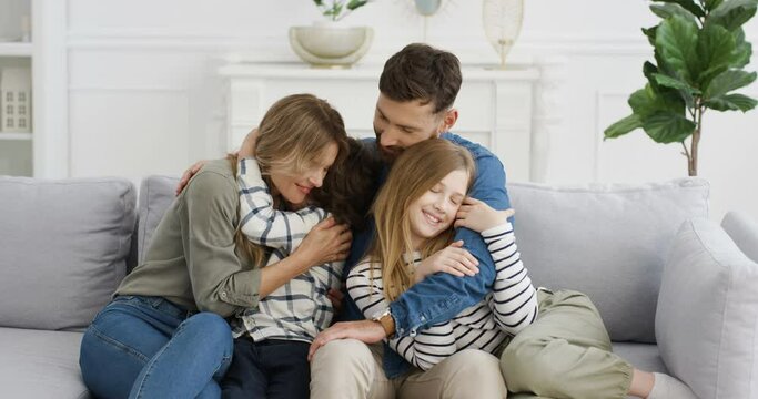 Portrait of young happy Caucasian parents and children sitting on sofa in hugs and smiling cheerfully to camera. Joyful kids with mother and father hagging on couch in living room. Warm embraces.