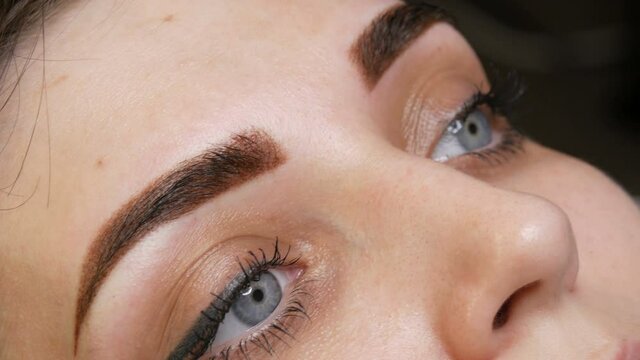 The face portrait of a beautiful young woman with blue eyes and long eyelashes after the procedure of permanent make up by microblading with eyebrow tattoo lies on a couch in a beauty salon close up.