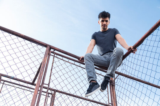 Hispanic young man on rooftop sitting watching sunset - man on top of old metal mesh frame with blue sky in background - urban style