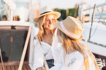 Two beautiful blonde girls friends mother and daughter in white and straw hats on the yacht at the pier