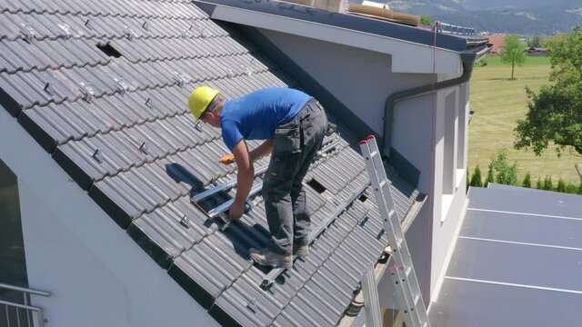 Young Caucasian man solar panel roof installer wearing yellow hard hat and blue shirt standing on steep roof of countryside residential home rolling orange wire on spool, static profile