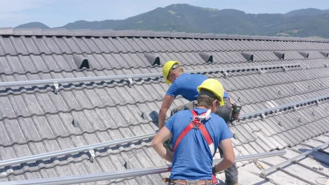 Two young Caucasian male solar power roof installers wearing yellow industrial hard hats walking on residential rooftop with mountain range in background on sunny day, handheld close up pan