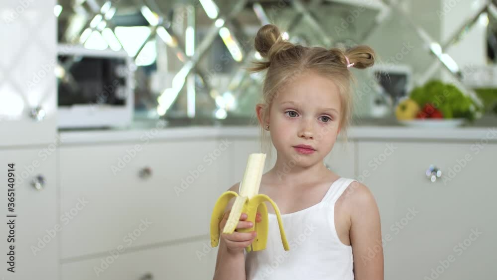 Wall mural Child Eating Food. Little Girl Eating Banana At Kitchen. Portrait Of Cute Kid With Fruit In Hand 