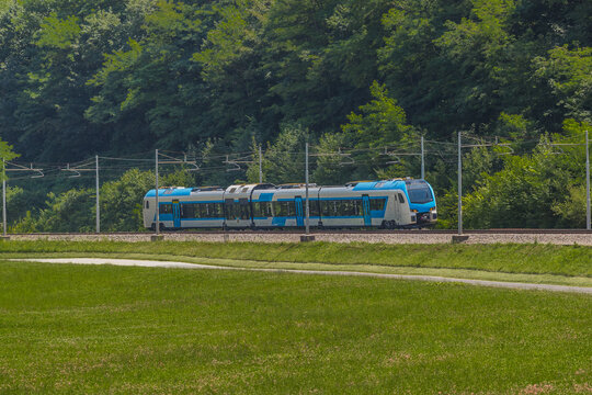 Modern and new passenger swiss made Train in white and blue color is traveling on a dual track railway line in the rural setting on a sunny hot day.