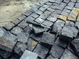 Natural granite stone is laid on a dry sand-cement base with a rubber mallet. The hammer tool lies among the gray-black cobblestones, close-up