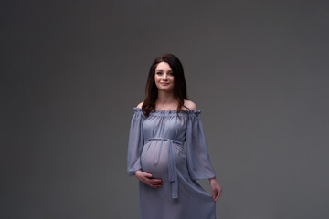 A young pregnant woman in a dress stands on a gray background.