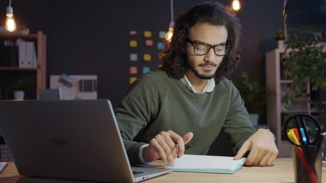 Mixed race man is using laptop computer writing information in notebook in dark office at night working overtime. People, business and lifestyle concept.