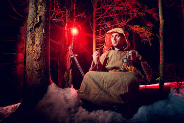 Naklejka na ściany i meble Medieval monk in canvas sackcloth robe praying in dark forest with snow and red light on winter night. Fantasy or fairy tale about wandering monk. Story about the forces of good and evil in world
