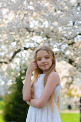 Beautiful blue-eyed girl with long blond hair in a white dress walking in the flower garden. summer bright, emotional photo.