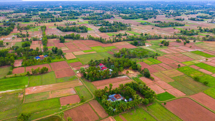 A top down aerial view of a small country town with traditional houses with orange roofs, a red dirt road, rice fields, and palm trees in the jungle in Cambodia.