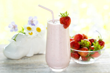 A glass of strawberry milkshake sits on the edge of a light wooden table