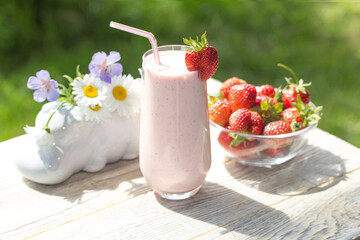 A glass of strawberry milkshake sits on the edge of a light wooden table