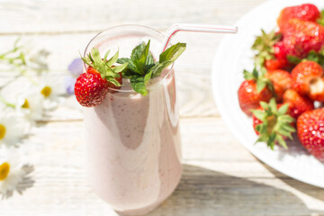 On a Sunny day, on a light wooden background, there is a glass glass with a strawberry milkshake , a white plate with strawberries and scattered daisies