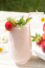 On a Sunny day, on a light wooden background, there is a glass glass with a strawberry milkshake , a white plate with strawberries and scattered daisies