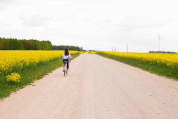 Fototapeta premium Young woman rides a bicycle on a road between a blooming rapeseed field. Yellow flowering rapeseed field. Bright spring bloom of rapeseed