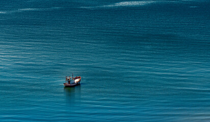 Aerial view of the sky, one lonely small local fishing boat on a vast and boundless sea. Traces of waves on the water surface and sparkling reflections, tiny grains from sunlight.