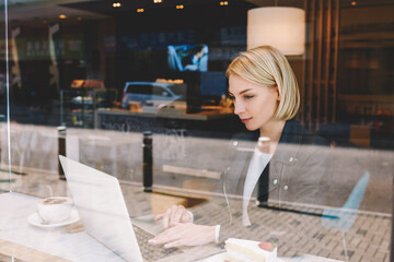 Young woman freelancer is ordering fly tickets on-line via laptop computer, while is sitting in modern hipster coffee shop interior. Smart female remote programmer is using net-book for distance work