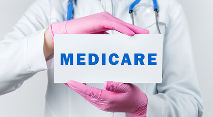 Cropped view of young female doctor in a white coat and pink sterile gloves holding a card with text MEDICARE. Medical concept.