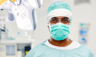 Fototapeta na wymiar medicine, surgery and people concept - indian male doctor or surgeon in mask, goggles and protective wear over operating room at hospital background
