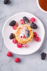 Cottage cheese pancakes and powdered sugar, curd fritters dessert with raspberry and blackberry berries in plate near to hot tea cup with lemon slice on stone concrete background, top view