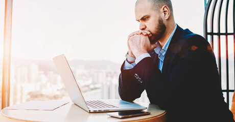 Man intelligent managing director is reading important e-mail letter on laptop computer, while is sitting in modern office interior. Worried economist is analyzing activities of  company via net-book