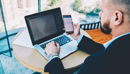 Cropped image of businessman dressed in formal wear making calculation on smarphone to checking financial reports received by mail on modern netbook with mock up area sitting in office interior