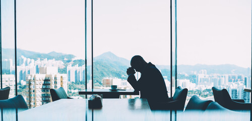Depressed male proud ceo tired from hard working day and unsuccessful business project sitting at table with cup of coffee in modern cafeteria with beautiful evening metropolis scenery