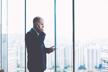 Man owner of successful company in the field of real estate trade is having serious mobile phone conversation with unsatisfied customer, while is standing near window with view of metropolitan city