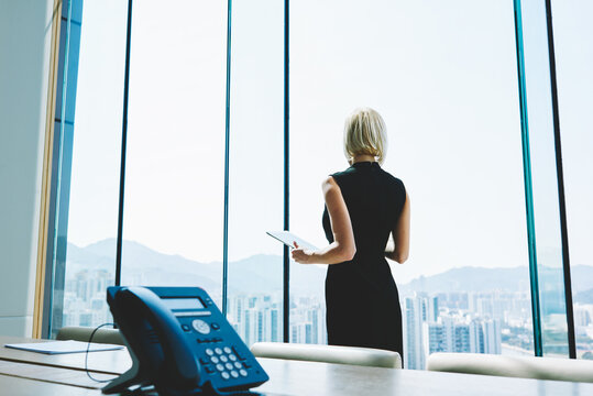 Woman proud CEO with digital tablet in hands thoughtful is looking into the office window, while is standing in skyscraper interior in big conference room with table. Copy space for advertising text
