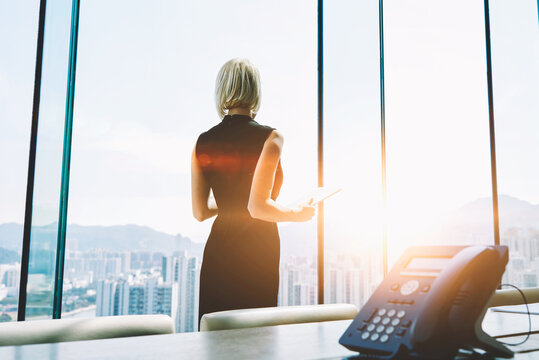 Back view of a businesswoman with digital tablet in hands is watching in office window on a Hong Kong city view and thinking about future her company. Female manager is using touch pad during work day