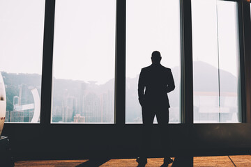 Fototapeta premium Back view of male financial manager dressed in formal wear looking out of window and thinking on developing company and business project standing in loft interior.Black silhouette of banker in office