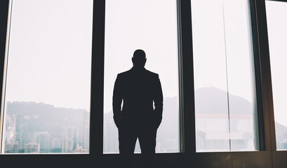 Fototapeta na wymiar Back view of male proud ceo dressed in elegant wear looking out of window while thinking on working issues after hard day.Silhouette of businessman standing indoors in office of own company