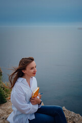 Attractive woman is sitting on the edge of the mountain with a sea view, reading a book.