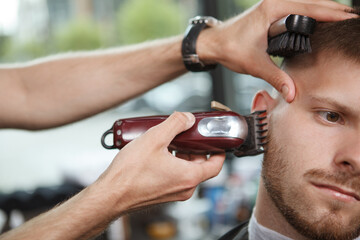 Cropped close up of unrecognizable barber using electric clipper, shaving beard of male client