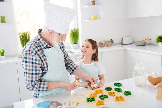 Nice work sweety. Photo of little pretty girl granddaughter help grandpa chef cap forming dough colorful forms baking cookies cake together free time weekend home kitchen indoors