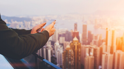 Closely of man successful broker is using mobile phone,while is standing on building roof against view of Hong Kong city with tall skyscrapers. Hipster guy is connecting to internet via cell telephone