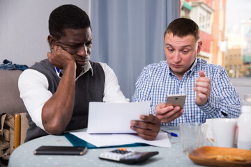 Upset interracial men using mobile in home interior, filling up documents