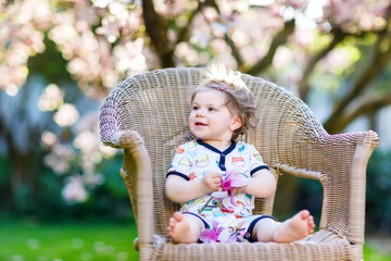 Cute little baby girl sitting on big chair in garden. Beautiful happy smiling toddler with blooming pink magnolia tree on background. Healthy child enjoying spring season.
