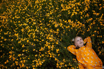 A girl in a yellow dress lies on the grass with yellow flowers and smiles.