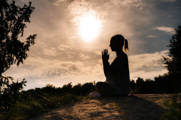 Silhouette of young yogini woman meditating in lotus position with closed eyes. Relaxed lady is performing namaste yoga pose background of sun outdoor on park outside city evening during sunset.