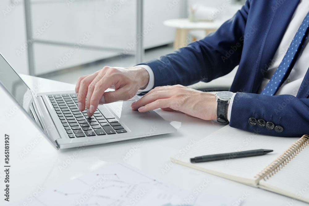 Aufkleber Close-up of unrecognizable businessman in jacket sitting at desk with notepad and typing on laptop in office
