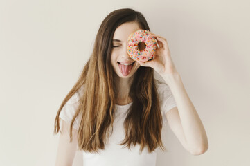 Portrait of cheerful young woman with funny emotion holding a pink donut with sprinkles near her eye