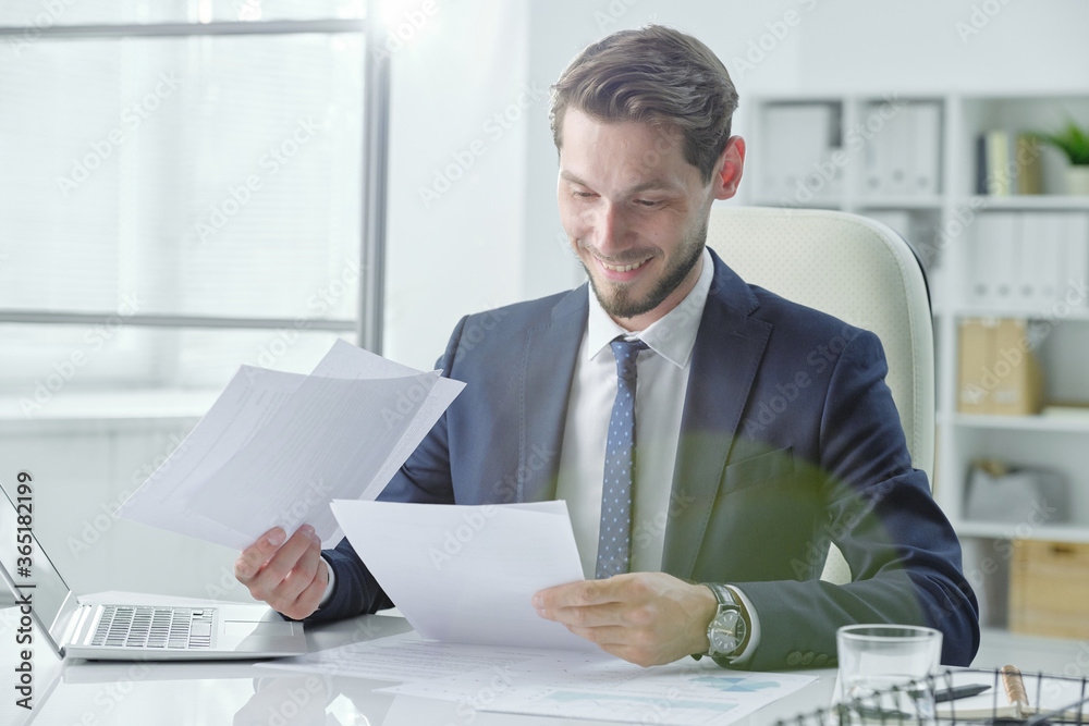 Poster satisfied young analyst sitting at desk and working with documents in office