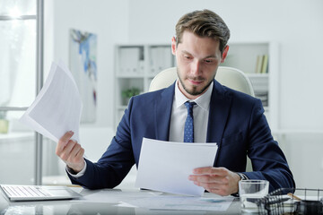 Busy young manager in formal suit sitting at desk and examining business papers in office